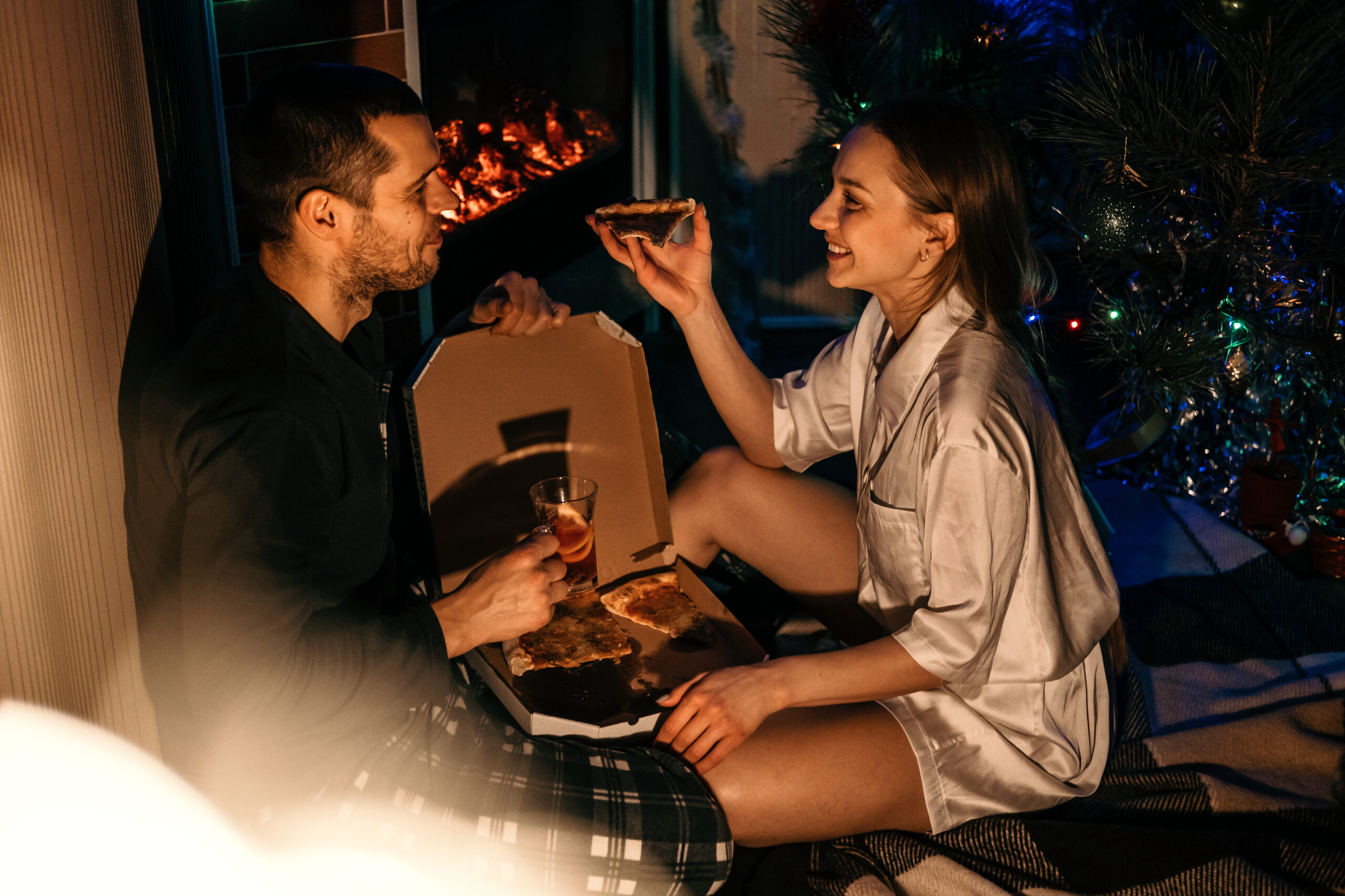 Valentines day home date ideas Save Money Budget Tips. Young couple in love celebrating Valentines day at home. Romantic couple in pajamas eating pizza near fireplace.