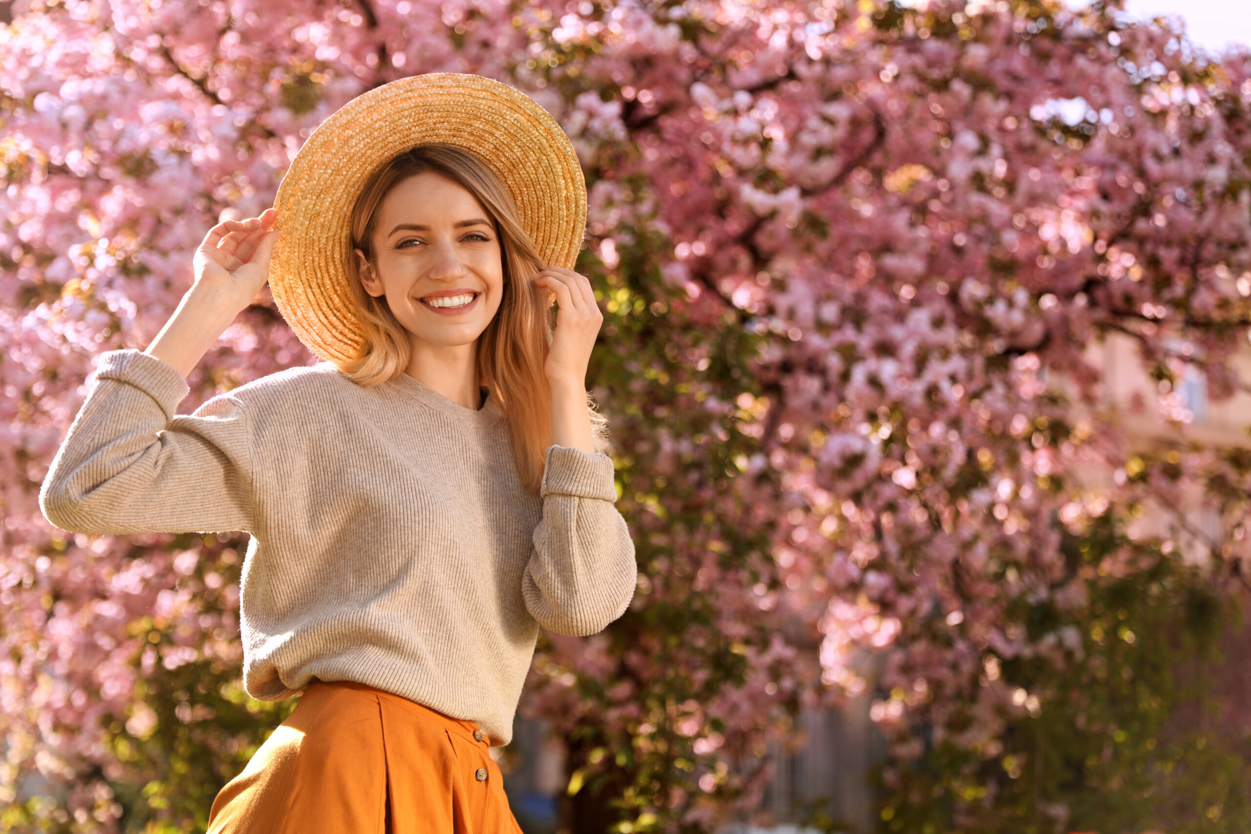 A young woman wearing a stylish outfit smiles as she stands in front of a cherry blossom tree